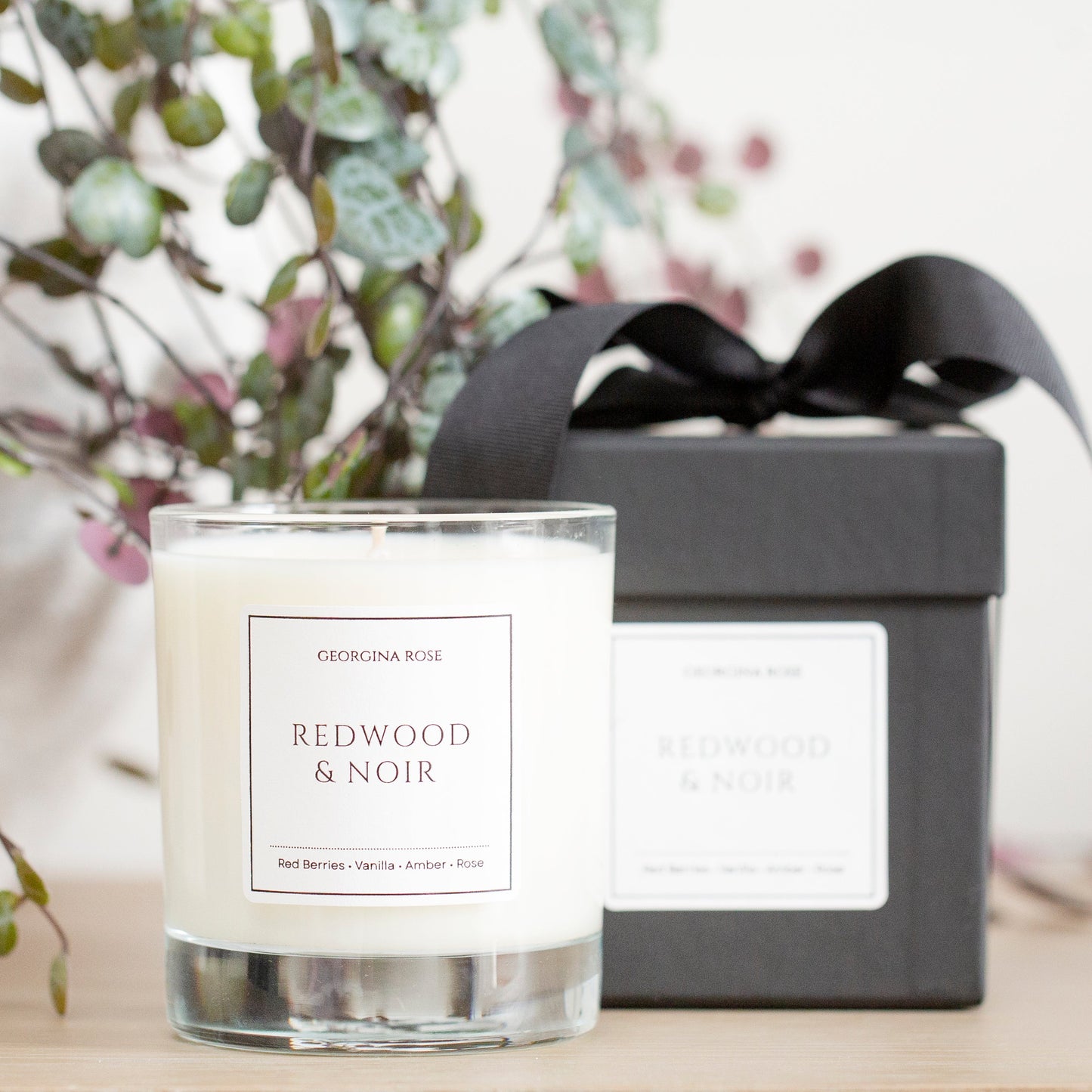 Candle labeled 'Redwood & Noir' with a black gift box and decorative plant in the background.